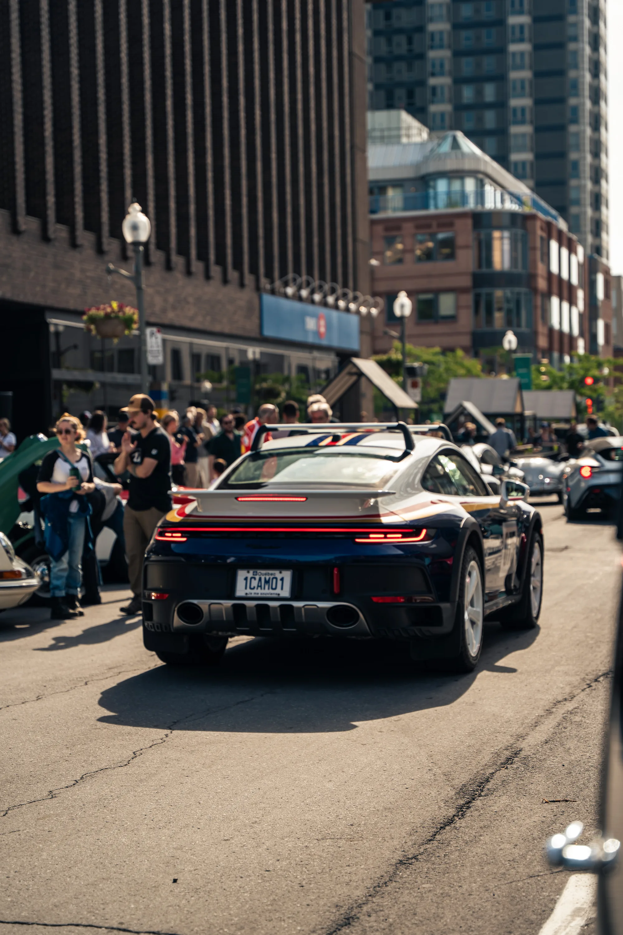A Porsche 911 in racing livery pulls away down the street as the crowd watches from the curb.