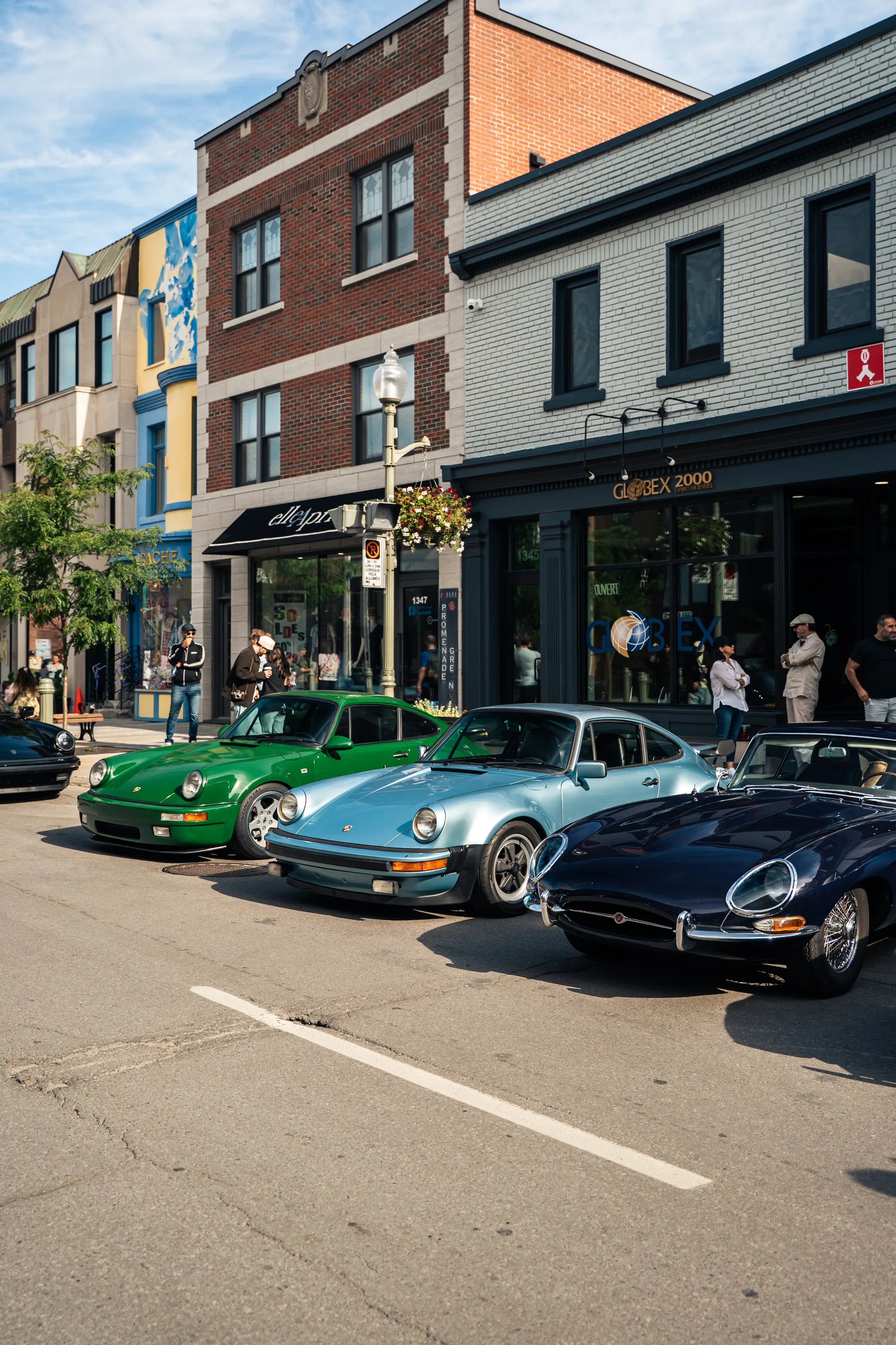 A green Porsche 911, a pale blue 911 and a dark blue Jaguar E-Type nose in at the curb in front of the shops.