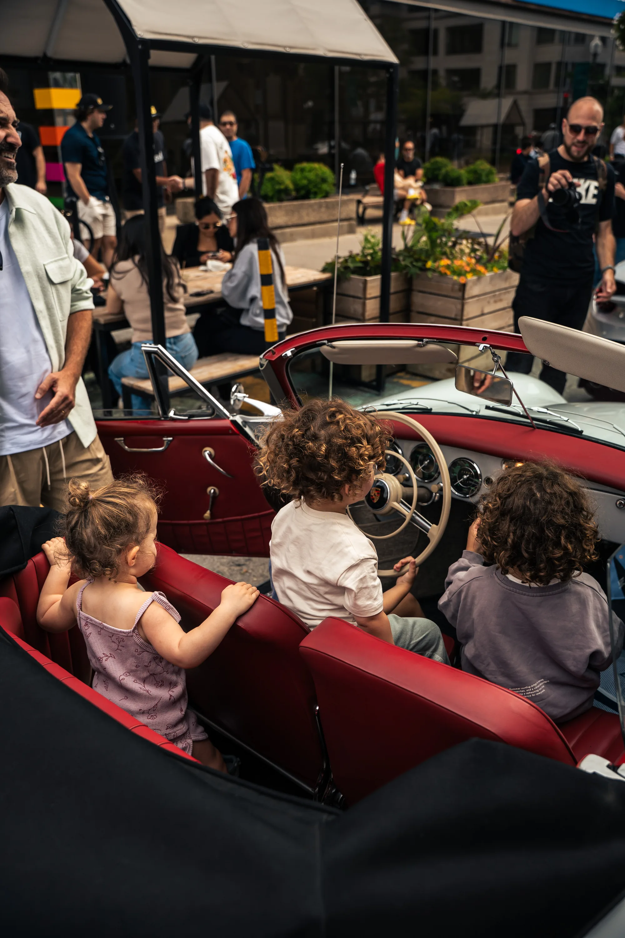 Three small children in the red leather interior of a cream convertible, one of them at the wheel.